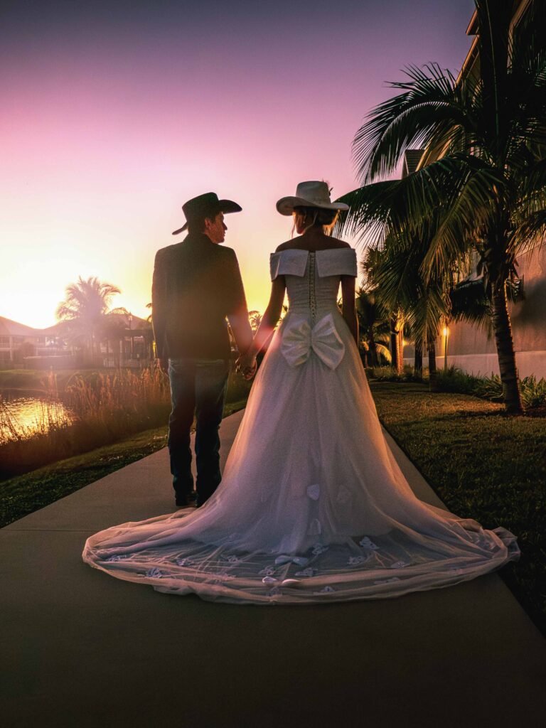 Newlyweds in elegant western wedding attire share an intimate moment walking through tropical gardens at sunset